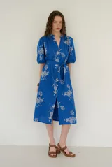 Vestido midi azul con estampado floral blanco, cuello en V, mangas cortas abullonadas y cierre frontal de botones. Incluye un lazo ajustable para la cintura.