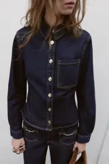 Camisa entallada de denim azul oscuro con cuello solapa, manga larga y cierre frontal con botones metálicos. Presenta un bolsillo de plastrón en el pecho y costuras a contratono.