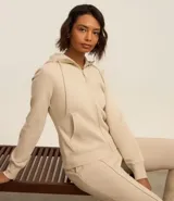 Campera deportiva color beige con cierre frontal de cremallera, capucha ajustable con cordones, mangas largas y bolsillos frontales tipo canguro. Confeccionada en tejido de algodón y poliéster.
