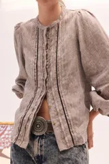 Blusa de lino color beige, de cuello redondo y manga larga abullonada. Cuenta con detalles de pliegues y ribetes oscuros en la pechera.