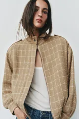 Chaqueta bomber de color beige con diseño a cuadros blancos, cuello subido, manga larga con trabillas en los hombros, bolsillos delanteros y bajo con elástico. Cierre frontal con cremallera y botones a presión.