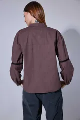 Camisa blanca de algodón con pechera decorada con alforzas y puntillas. Presenta cierre frontal con botones, mangas tres cuartos con frunces y detalles de puntilla, y un diseño de corte holgado con ruedo redondeado.