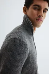 Buzo de punto acanalado en color gris, con cuello elevado y cierre frontal parcial. Presenta un diseño de corte estándar con puños y bajo acanalados.