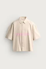 Sobrecamisa boxy fit de algodón color beige, de manga corta, con cuello solapa, cierre frontal de botones y aplicación de texto en parches rosas a contraste en el delantero.
