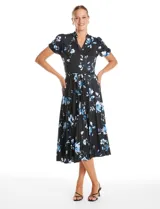 Vestido midi de seda fría con estampado floral en tonos azules sobre fondo negro. Presenta escote en V con cierre frontal de botones, mangas cortas con terminación fruncida, corte en la cintura con lazo de ajuste y falda plisada.