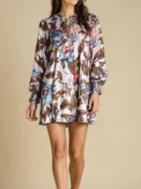 Vestido corto blanco con estampado floral en tonos marrones, azules y rosas, escote en V y mangas largas con puños elásticos.