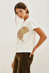 Remera de algodón color blanco con cuello redondo y mangas cortas con dobladillo. Presenta un bordado detallado de una mariposa en tonos dorados en la parte frontal.