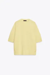 Jersey de punto color amarillo pálido, con cuello redondo y mangas cortas. Presenta un calce oversize y aberturas caladas en los laterales inferiores.