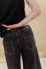 Pantalón de jean gris oscuro, estilo sastrero de tiro medio bajo y corte ancho (wide leg). Presenta costuras marcadas en el frente y un cinturón trenzado marrón con hebilla metálica.