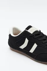 Zapatilla urbana de perfil bajo con diseño retro, confeccionada en poliuretano con detalles en textura tipo gamuza. Presenta puntera redondeada, cordones clásicos y suela de goma color caramelo.