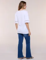 Remera blanca de silueta recta y algodón suave, con escote redondo y pequeña abertura central tipo gota. Presenta mangas de largo intermedio con aplicación horizontal de puntilla calada al tono y ruedo recto con tajos laterales.
