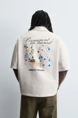 Camisa de corte relajado confeccionada en algodón, con cuello bowling y manga corta. Presenta cierre frontal con botones y detalle de texto estampado en el pecho.