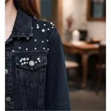 Campera de jean color negro con apliques de perlas en la parte superior frontal. Presenta cuello clásico, cierre frontal con botones metálicos y dos bolsillos en el pecho.