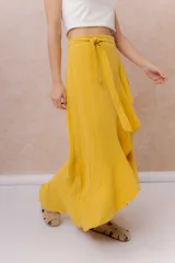 Falda larga envolvente de color mostaza, con diseño asimétrico, lazo ajustable en la cintura y volados en cascada.
