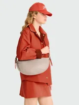 Bolso tipo crossbody de diseño semicircular, color gris claro, con correa ajustable y cierre superior.
