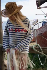 Sweater de tejido de punto a rayas horizontales en color crudo y azul marino, con cuello redondo y mangas largas. Presenta la inscripción 'LA BRAVA' bordada en el frente en color rojo y flecos largos en el ruedo.