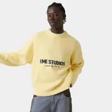 Suéter de punto grueso color amarillo, con corte cuadrado y ligeramente corto. Presenta cuello redondo, mangas largas con puños ajustados y el logo de la marca Eme Studios junto a la frase 'curated by ben ice' en jacquard oscuro en el centro del pecho.