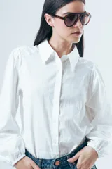 Camisa blanca de manga larga con diseño de rayas verticales sutiles en la misma tela. Presenta cuello clásico, cierre frontal con botones y mangas abullonadas con puños elásticos.