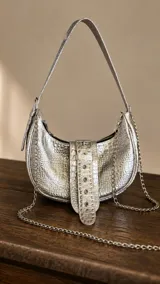 Cartera tipo bandolera y morral plateada con textura de cocodrilo, diseño curvo, asa superior y correa de cadena metálica. Cuenta con una tira frontal con hebilla y tachas decorativas.