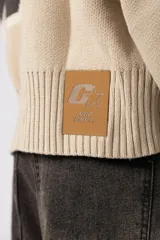 Campera tejida de punto con cierre frontal completo y cuello polo. Presenta un diseño de franja horizontal ancha en color crema con estampado gráfico oscuro sobre una base de color marrón. Posee puños y ruedo acanalados, con un pequeño parche de marca en el puño izquierdo.