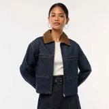 Chaqueta vaquera de color azul oscuro, corte corto y holgado, con cierre frontal de cremallera y dos bolsillos grandes con solapa en el frente. Posee cuello de pana marrón.