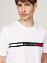 Remera blanca de algodón con cuello redondo y manga corta. Presenta una banda horizontal con los colores distintivos de Tommy Hilfiger y el logo bordado en el pecho.