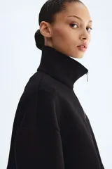 Campera negra de efecto neopreno, con cuello chimenea, cierre frontal completo y hombros caídos. Presenta un diseño holgado con pliegues en el bajo y mangas largas.