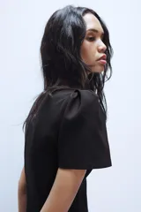Vestido corto de color negro con cuello redondo y mangas cortas abullonadas. Presenta un diseño entallado con costuras frontales y cierre de cremallera oculto en la espalda.