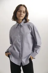 Camisa de corte oversize en color azul claro, confeccionada en algodón. Presenta cuello clásico, cierre frontal con botones, mangas largas con puños abotonados y un bolsillo aplicado en el pecho.