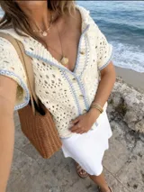Blusa tejida en crochet de color crema con detalles de ribetes en color celeste. Presenta un diseño calado, cuello solapa, cierre frontal con botones y mangas cortas.
