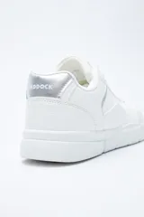 Zapatilla urbana blanca de mujer, con silueta moderna y líneas limpias. Presenta detalles en poliuretano liso y un panel plateado en el talón. La suela es de perfil recto y firme.