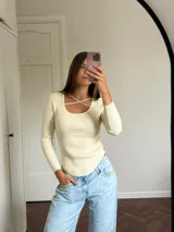 Polera de punto acanalado color blanco, con escote redondo y detalle de tiras cruzadas en el pecho, de manga larga y corte ajustado con ruedo curvo.