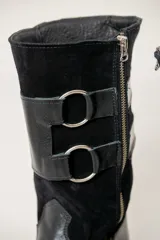 Bota de caña media combinada en cuero liso y gamuza, color negro. Presenta un diseño con doble hebilla lateral metálica y cierre de cremallera vertical en el lateral interno. Cuenta con suela plana y tacón bajo.