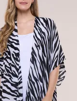 Kimono confeccionado en gasa ligera de poliéster, con diseño estampado animal print de cebra en color blanco y negro. Presenta silueta amplia, mangas cortas integradas y ruedo asimétrico con detalles de borlas en las puntas.