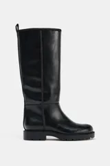 Bota de caña alta estilo plano, confeccionada en material sintético negro con acabado liso. Presenta detalle de pespuntes verticales en el cuerpo, punta redonda, suela track dentada y tirador trasero para facilitar el calce.