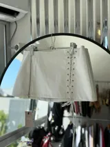 Minifalda blanca de cuero sintético, corte evasé, con cierre lateral y detalle de cordones cruzados en el frente.