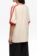 Remera oversize de color crudo con escote en V y cuello en rib rojo, mangas cortas con rib rojo y una franja roja en los hombros. Cuenta con un estampado frontal de bailarinas y detalles en relieve.