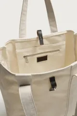 Bolso tipo shopper de tejido color crudo, con doble asa de hombro y detalles en cuero negro. Cuenta con compartimento principal con cierre de tira y botón a presión.