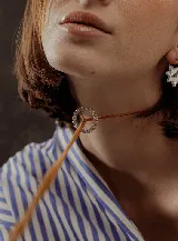 Choker marrón de gamuza con dije de doble aro entrelazado, uno liso y otro con incrustaciones de strass. Los extremos del choker tienen detalles de cuentas doradas.