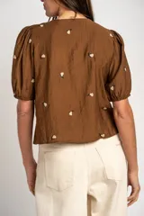 Blusa de viscosa color marrón, con escote en V, mangas cortas abullonadas y cierre frontal con botones. Cuenta con pequeños detalles bordados distribuidos por la prenda.