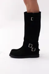 Bota de caña alta color marrón chocolate, confeccionada en gamuza. Presenta dos correas desmontables con hebillas metálicas plateadas.