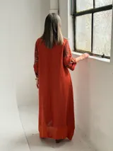 Vestido largo color terracota con diseño asimétrico que incluye un lateral con estampado floral grande. Presenta mangas tres cuartos, escote redondo y un bolsillo aplicado en el frente.