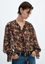 Blusa de gasa semitransparente con estampado floral en tonos rosa, verde y negro. Tiene cuello en V con lazo, cierre frontal con botones, mangas largas abullonadas con puños elásticos y corte recto.