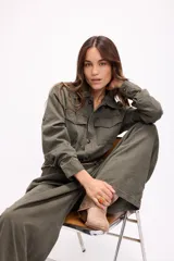 Mono de estilo utilitario color beige, con cierre frontal de botones, bolsillos en el pecho y cintura entallada con detalle de cadenas decorativas. Presenta mangas largas y piernas de corte recto y holgado.