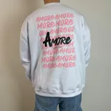 Buzo de cuello redondo y corte oversize, color blanco, con estampa gráfica en la espalda que repite la palabra 'Amore' en tonos rosados y un diseño central destacado en negro.