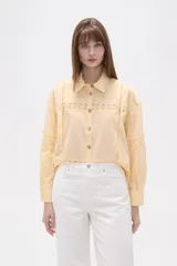 Camisa de algodón color amarillo claro, de corte holgado y diseño con detalles de encaje calado en el frente y las mangas. Presenta cuello camisero, cierre frontal con botones y mangas largas con puños rectos.