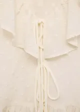 Blusa de gasa color crema con textura de pequeños puntos en relieve, cuello en pico con volantes, mangas largas abullonadas con puños acampanados y cierre frontal mediante lazos.
