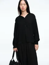 Camisa negra de corte holgado con sutiles destellos brillantes en la textura. Presenta cuello camisero, cierre frontal parcial con botones y mangas largas con puños estructurados.
