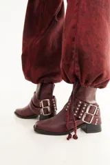 Bota de caña baja confeccionada en cuero con textura crocco, color bordó. Presenta taco cuadrado, punta cuadrada y un accesorio desmontable con doble hebilla metálica y tachas decorativas.
