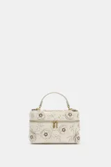 Bolso de mano formato vanity color crudo, con apliques de flores en relieve y hojas en el cuerpo. Cuenta con asa corta superior y cierre con cremallera doble dorada.